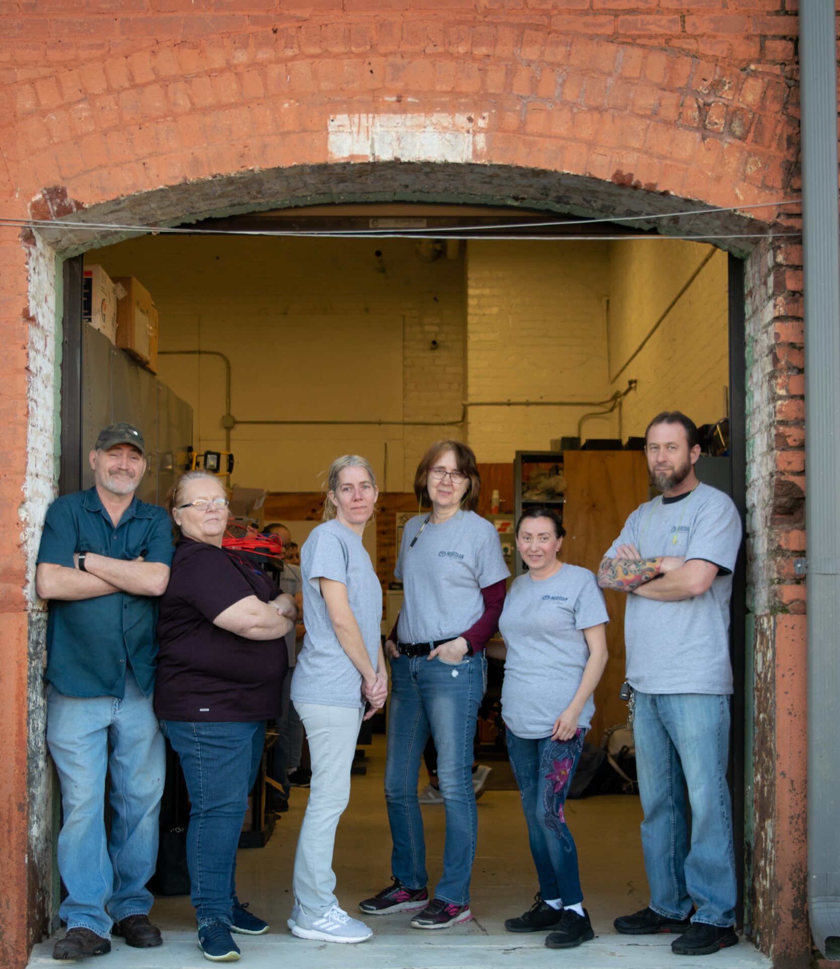 Seven adults standing in a wide brick doorway, posing for the camera in a warehouse, wearing casual gray shirts and jeans.