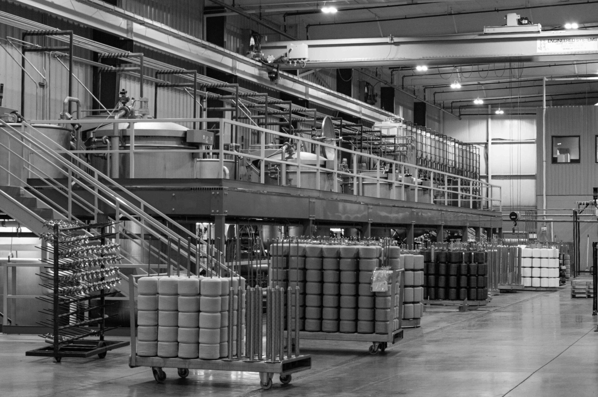 Industrial manufacturing floor with large stainless machinery, catwalks, and staircases; rows of stacked cylindrical foam rolls on carts under a raised platform.